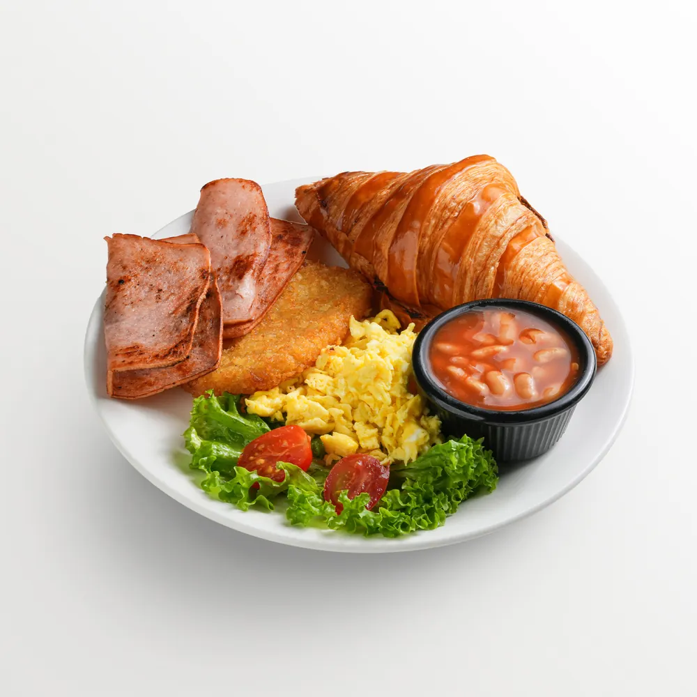 White background food photography of a big breakfast plate — turkey ham, butter croissant, hash brown, scrambled egg, baked beans and salad